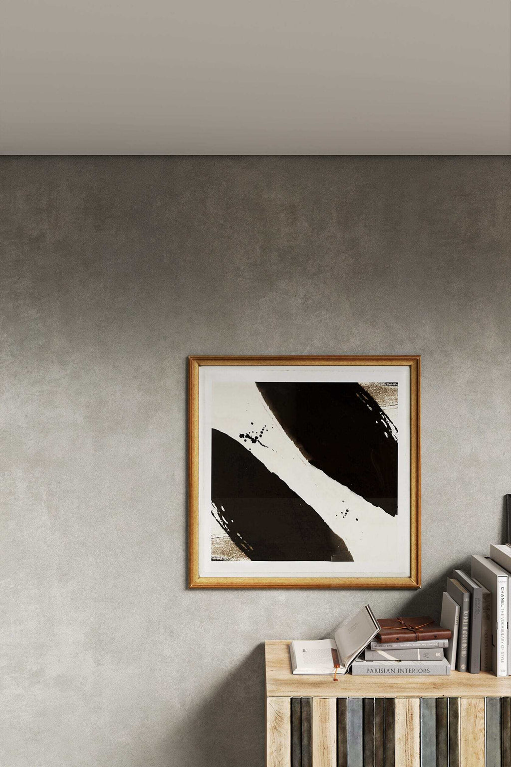 A framed abstract wall art print with bold black brushstrokes on a white background, enclosed in a light wood frame and displayed on a textured gray wall above a wooden shelf with books.